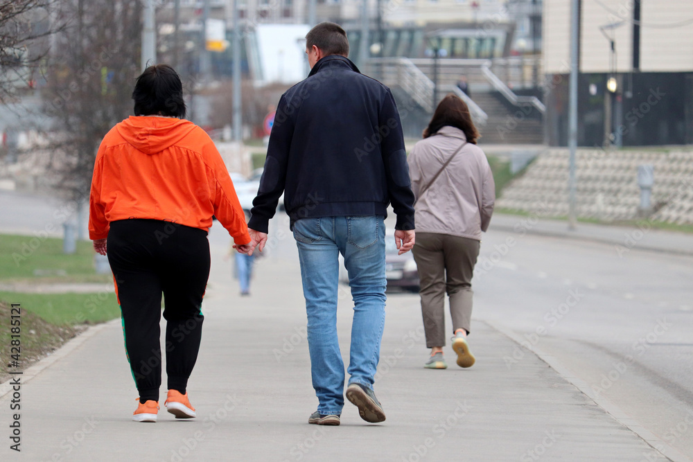 Fat woman in gym clothes and man in jeans walking along a city street. Couple in love holding hands, spring fashion