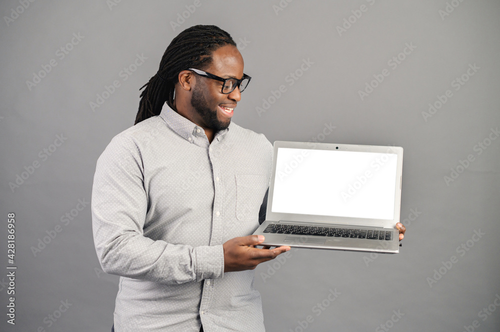 Mixed-race black man with dreadlocks standing isolated over grey background, presenting new application, showing laptop screen with white blank space for advertisement, mockup image