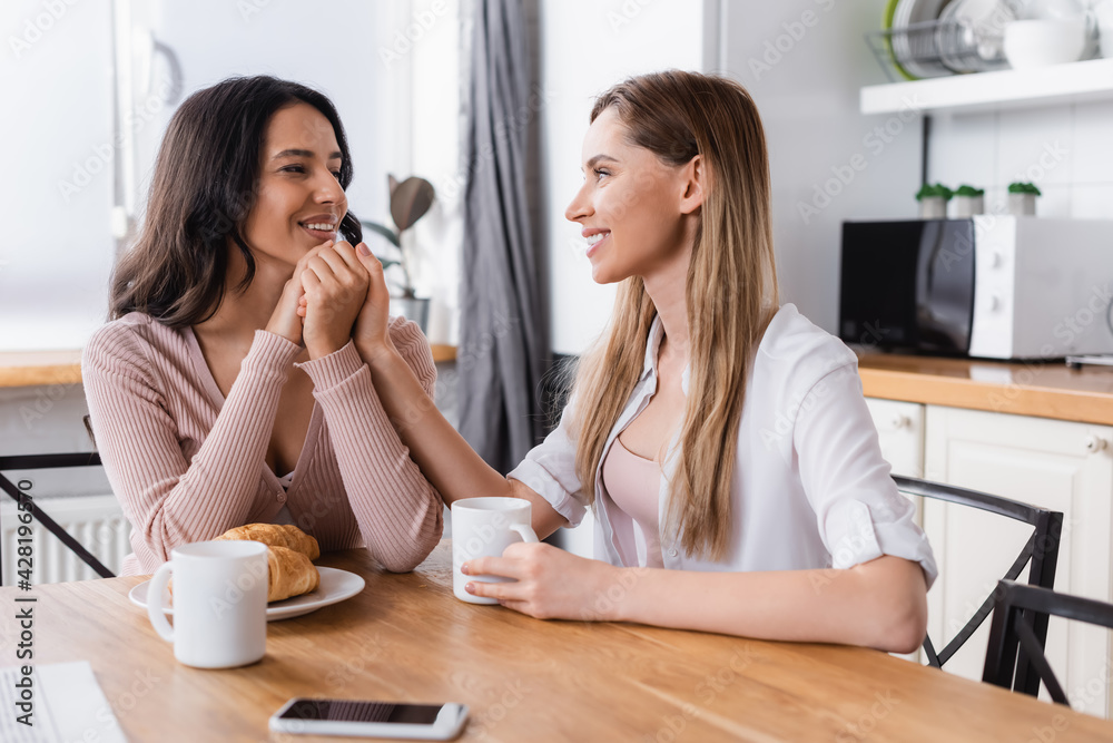 happy lesbian couple holding hands near breakfast and smartphone on kitchen table happy lesbian couple holding hands near breakfast and smartphone on kitchen table