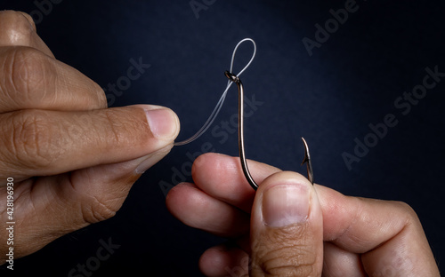 Fishing line and hook being handled by the hands of a brown man demonstrating how to thread the line correctly on a black and dark background. Used for fishing and family leisure.