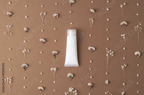 White tube of cosmetic cream with flowers on dark brown background. Flat lay