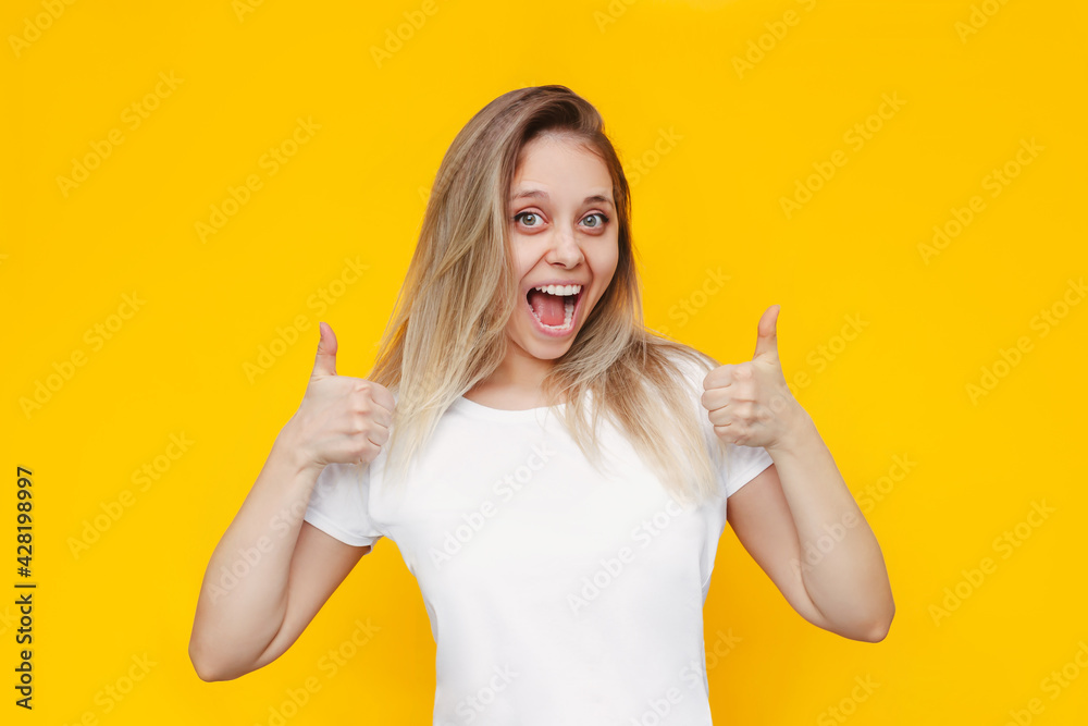 A young caucasian beautiful happy excited smiling blonde woman in a white t-shirt showing thumb up like gesture with two hands isolated on a bright color yellow background. Positive human emotions A young caucasian beautiful happy excited smiling blonde woman in a white t-shirt showing thumb up like gesture with two hands isolated on a bright color yellow background. Positive human emotions