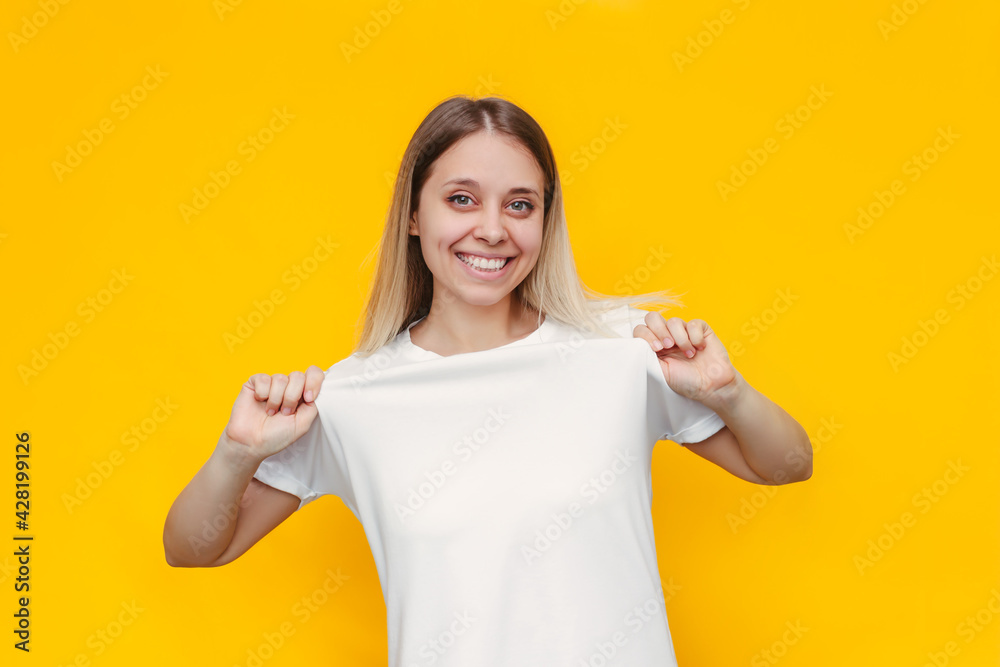 Portrait of a young caucasian happy smiling blonde woman demonstrates her white casual t-shirt, showing empty space for design and text with copyspace isolated on a bright color yellow background Portrait of a young caucasian happy smiling blonde woman demonstrates her white casual t-shirt, showing empty space for design and text with copyspace isolated on a bright color yellow background
