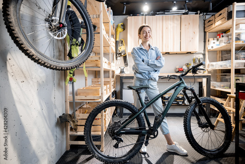 Portrait of a young handywoman standing with a bicycle in the workshop or garage at home. Bicycle repairing and diy concept