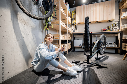 Young relaxed handywoman sitting with phone while repairing her bicycle in the well equipped workshop at home. DIY concept