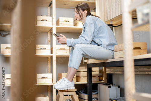 Portrait of a young handywoman sitting with smart phone on the workbench in the well equipped workshop at home. DIY conceept