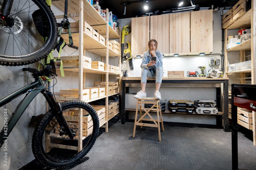 Well equipped home workshop with bicycles, wooden shelves and woman sitting on a workbench - obrazy, fototapety, plakaty 