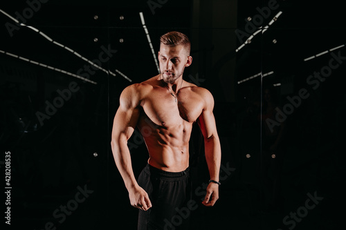 Athletic shirtless young male fitness model holds the dumbbell with light isolated on dark background.