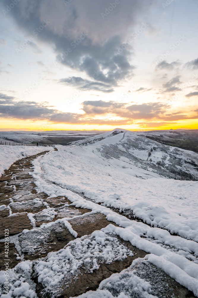 Icy snow covered winding stone footpath path way leading up mountain side as sun starts to come above horizon at sunrise winter morning. Perspective leading away to hill side with no people orange sky