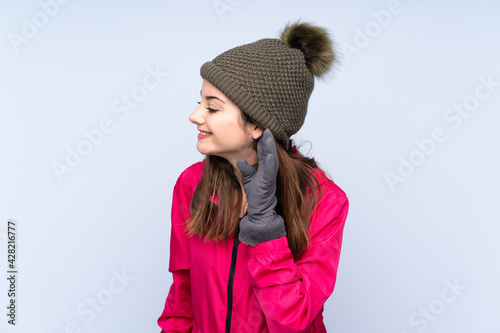 Young girl with winter hat isolated on blue background listening to something by putting hand on the ear