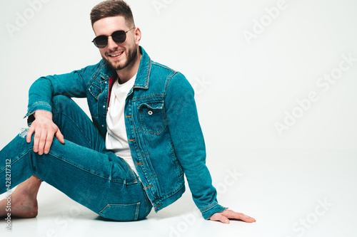 Portrait of handsome smiling stylish hipster lambersexual model.Man dressed in jacket and jeans clothes. Fashion male sitting on grey background in studio in sunglasses