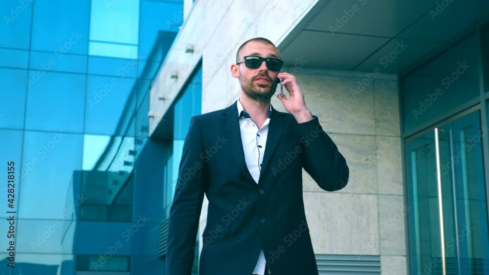 Businessman in sunglasses walking near office and talking on phone. Young happy business man going outdoor and speaking on cellphone. Portrait of guy having work conversation. Slow motion Close up Businessman in sunglasses walking near office and talking on phone. Young happy business man going outdoor and speaking on cellphone. Portrait of guy having work conversation. Slow motion Close up