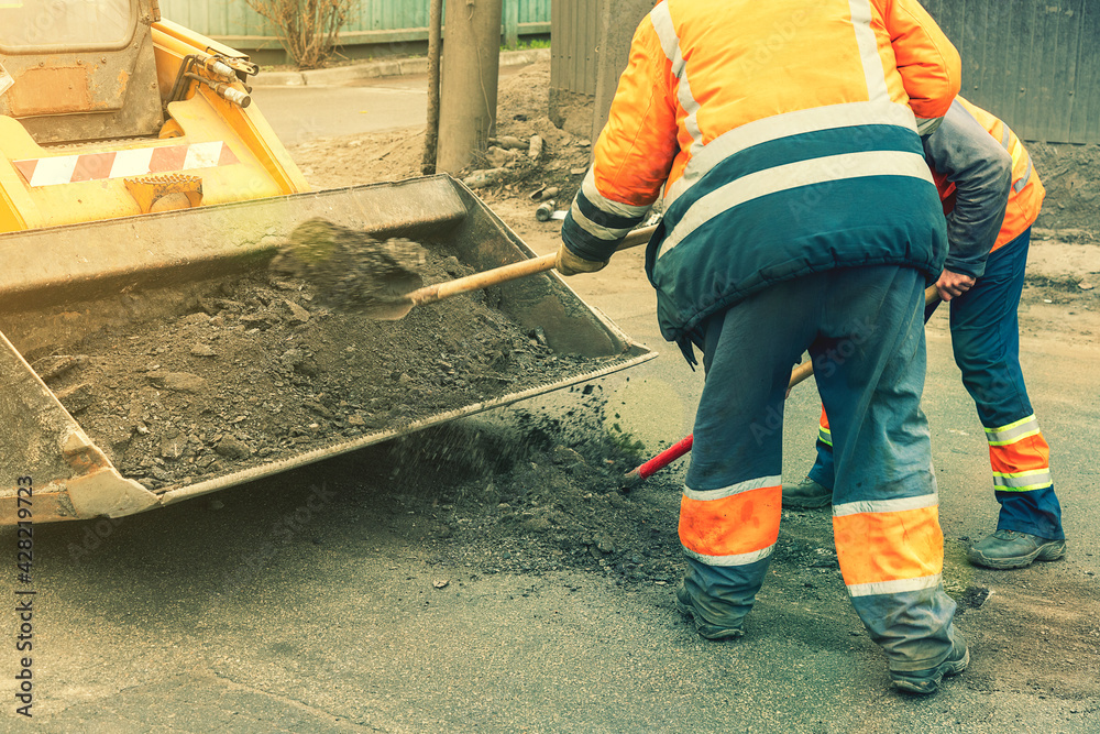 Road workers in bright orange reflective uniforms use shovels to scrape accumulated sand. maintenance of road and highway pavements. pothole repairing works. toned Road workers in bright orange reflective uniforms use shovels to scrape accumulated sand. maintenance of road and highway pavements. pothole repairing works. toned