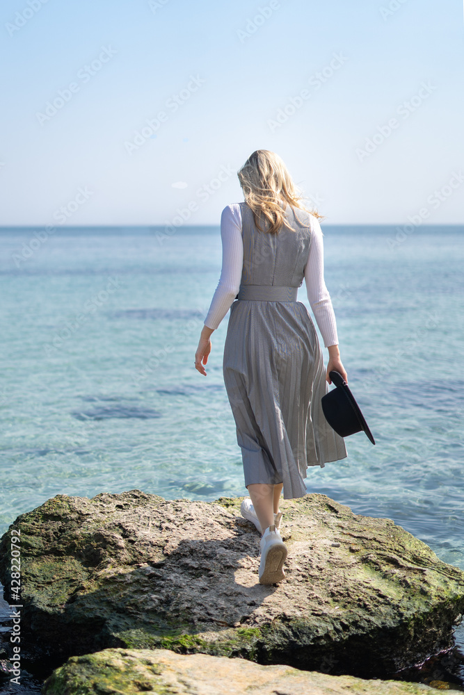 Stylish woman walking on the rocks at the beach near a clear blue sea water from behind holding a hat exploring the nature 