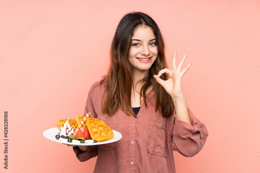 Young caucasian woman holding waffles isolated on pink background showing ok sign with fingers Young caucasian woman holding waffles isolated on pink background showing ok sign with fingers