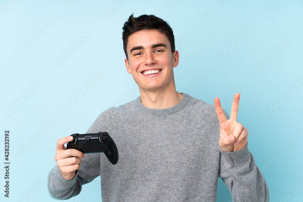 Teenager caucasian man playing with a video game controller isolated on blue background smiling and showing victory sign Teenager caucasian man playing with a video game controller isolated on blue background smiling and showing victory sign