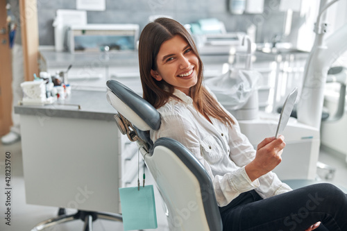 Happy young woman smiling checking out her perfect healthy teeth in the mirror  sitting in a dental chair at the dentist office