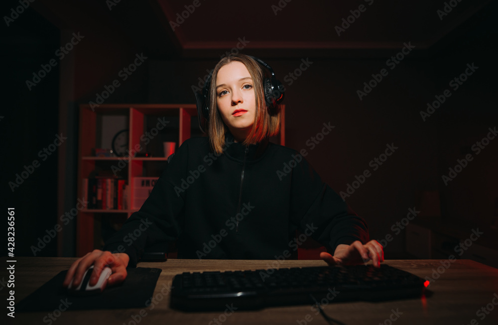 Attractive young woman in casual clothes sits at night at the computer in headphones and plays video games on the computer with a serious face. Attractive young woman in casual clothes sits at night at the computer in headphones and plays video games on the computer with a serious face.