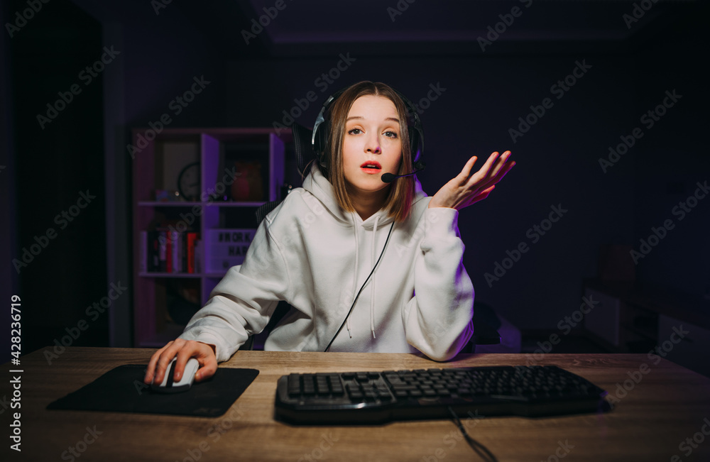 Surprised female gamer in white casual clothes and headset plays online computer games in the bedroom and looks at the screen with a dissatisfied face.
