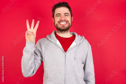 Young caucasian man wearing tracksuit over red background showing and pointing up with fingers number three while smiling confident and happy.