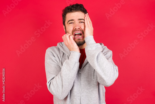 Doleful desperate crying Young caucasian man wearing tracksuit over red background, looks stressfully, frowns face, feels lonely and anxious