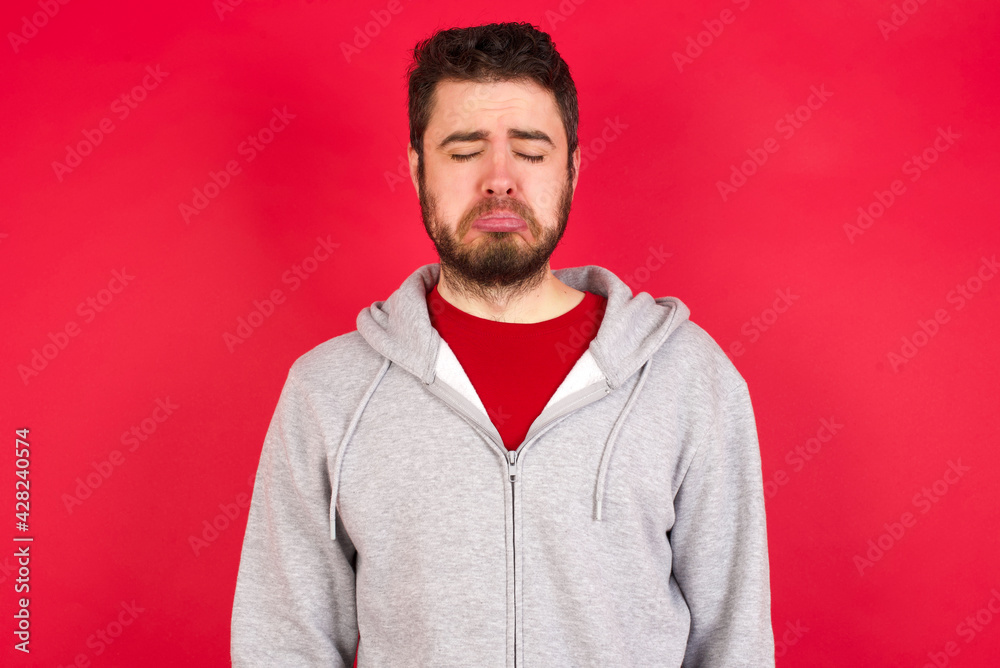Dismal gloomy rejected Young caucasian man wearing tracksuit over red background has problems and difficulties, curves lower lip and closes eyes in despair, being in depression Dismal gloomy rejected Young caucasian man wearing tracksuit over red background has problems and difficulties, curves lower lip and closes eyes in despair, being in depression