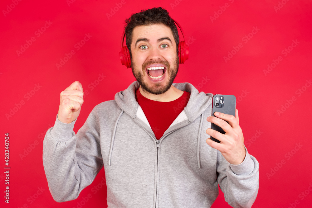Positive Young caucasian man wearing tracksuit over red background holds modern cell phone connected to headphones, clenches fist from good emotions, exclaims with joy, Positive Young caucasian man wearing tracksuit over red background holds modern cell phone connected to headphones, clenches fist from good emotions, exclaims with joy,
