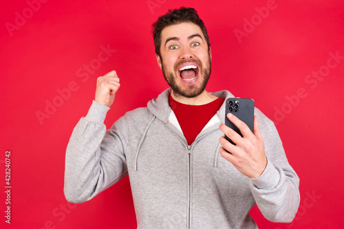 Portrait of Young caucasian man wearing tracksuit over red background holding in hands cell celebrating