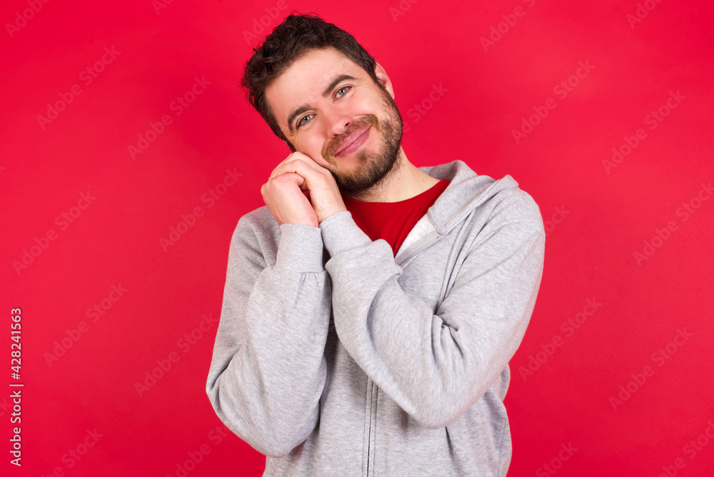 Happy Young caucasian man wearing tracksuit over red background keeps fists on cheeks smiles broadly and has positive expression being in good mood