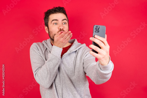 Young caucasian man wearing tracksuit over red background blows air kiss at camera of smartphone and takes selfie, sends mwah via online call. © Roquillo