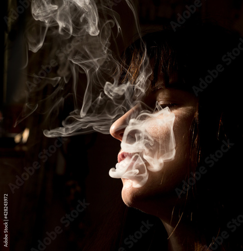 A large portrait of a girl in smoke. Cheek, nose, and hair in the sunlight. The woman blows white smoke out of her mouth. Dark Key