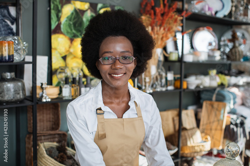 Close up of smiling confident African female worker in beige apron, posing at camera during work at decor shop. Stylish home decorations on the shelves