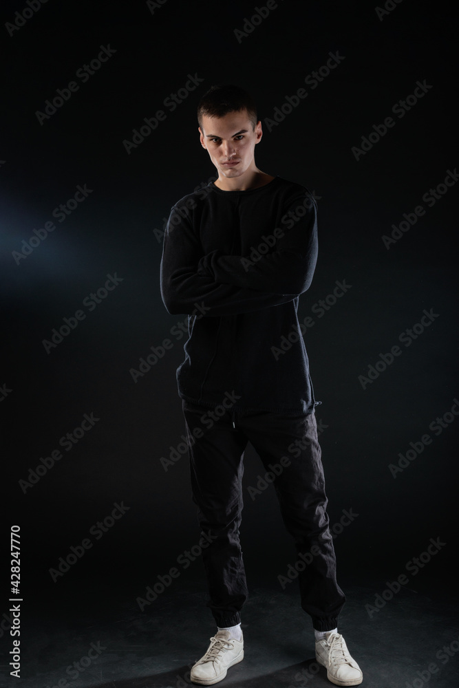 Full-length picture of an attractive sexy young male model posing isolated on a black background and wearing black blouse and black pants.