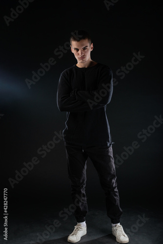 Full-length picture of an attractive sexy young male model posing isolated on a black background and wearing black blouse and black pants.