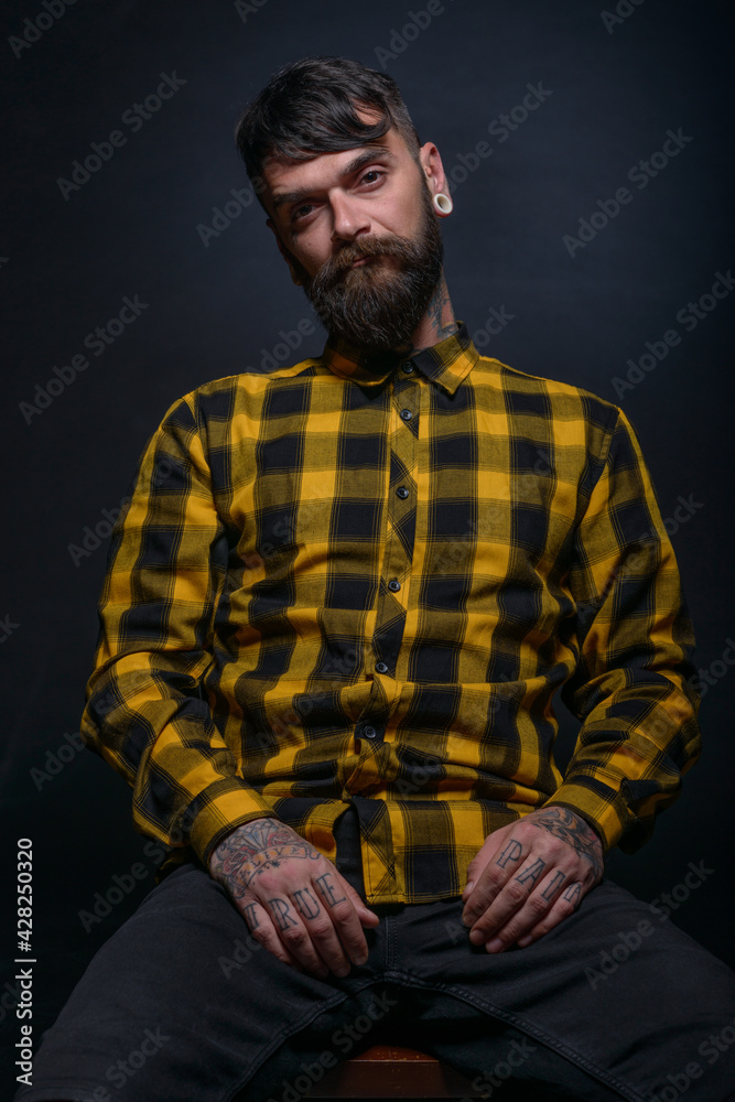 Tattooed man with a beard wearing checkered shirt is posing seated on a chair in a studio Tattooed man with a beard wearing checkered shirt is posing seated on a chair in a studio