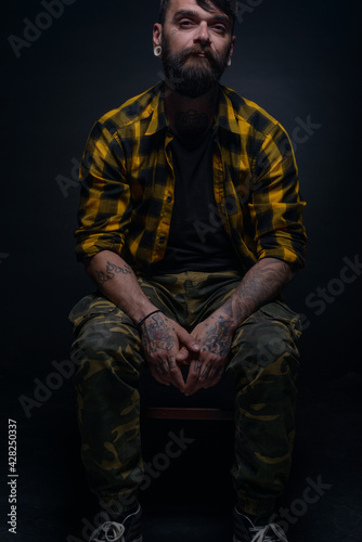 Handsome male model with tattoos and tartan shirt is sitting on a chair isolated on a black background