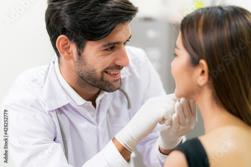 Male surgeon examining young asian female's face in modern clinic.