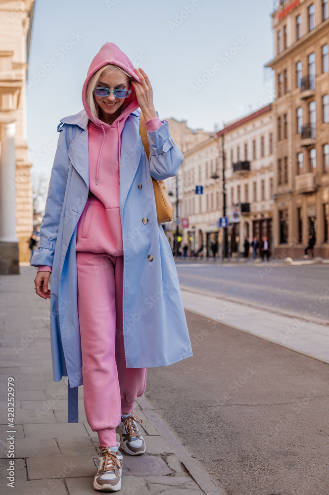 Happy smiling woman wearing trendy sport chic style outfit walking in city.  Streetstyle, street fashion conception. Blue trench coat, sunglasses, pink  hoodie, trousers, sneakers, yellow bag foto de Stock | Adobe Stock