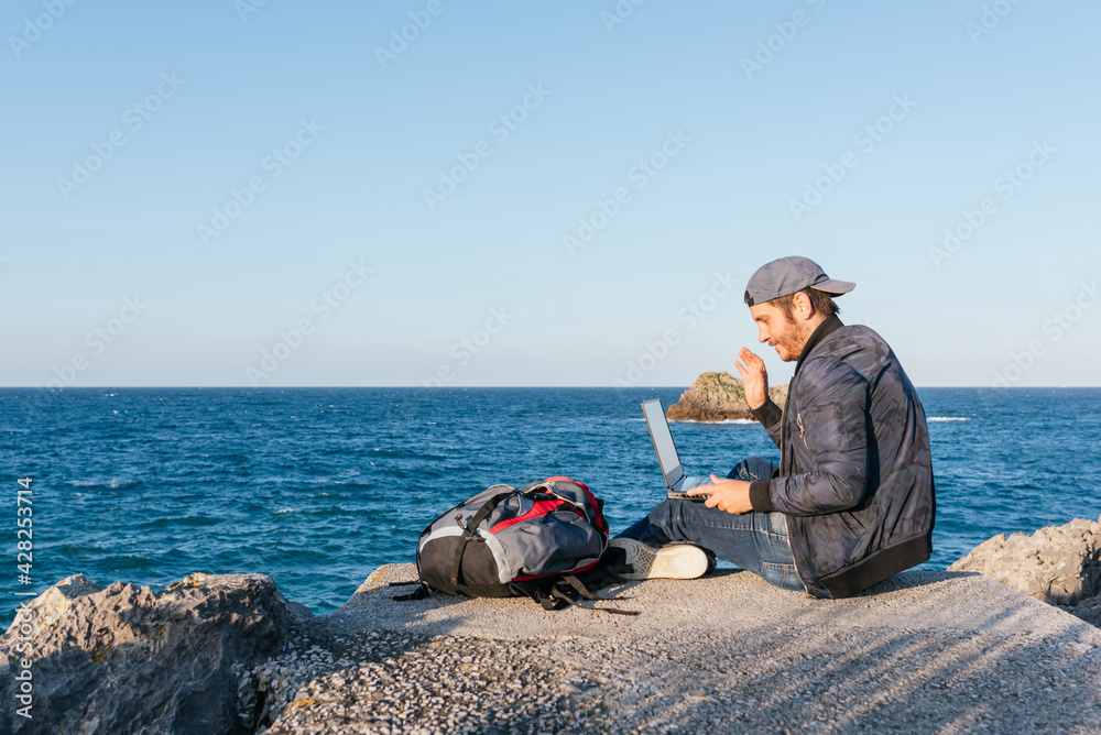 Backpacker man making a video call through his laptop with his family during his trip. Person using a computer in front of the sea.