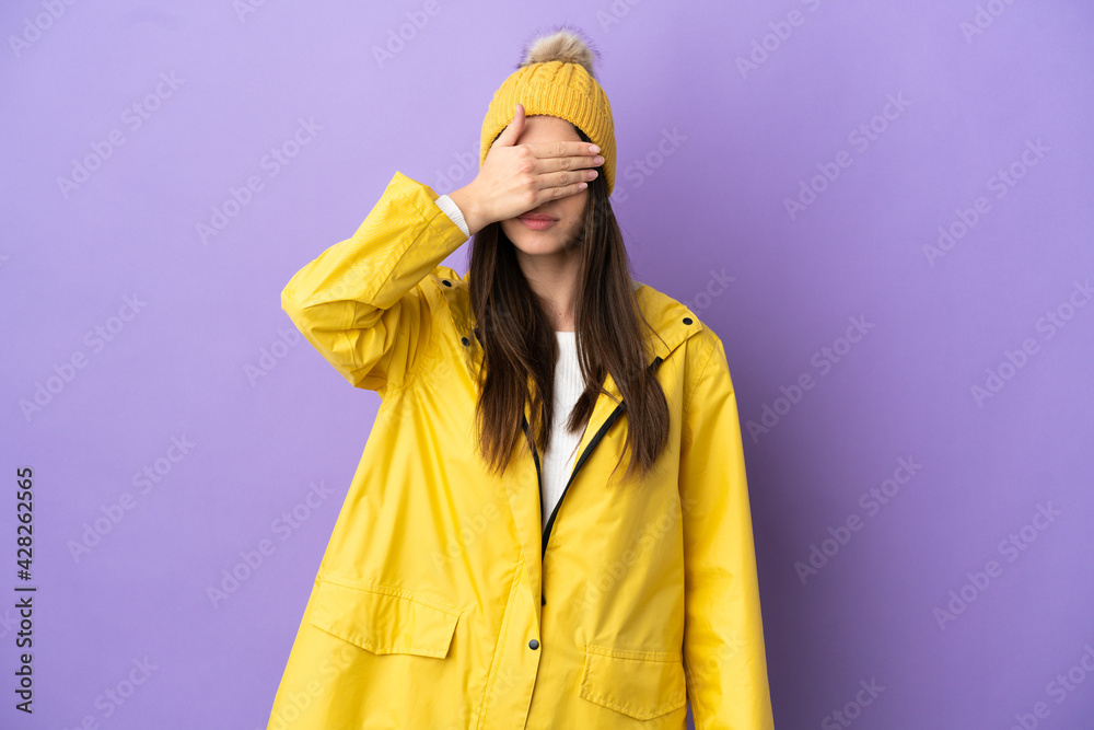 Young caucasian woman wearing a rainproof coat isolated on purple background covering eyes by hands. Do not want to see something Young caucasian woman wearing a rainproof coat isolated on purple background covering eyes by hands. Do not want to see something