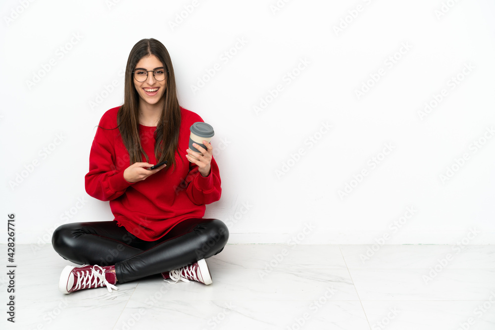 Young woman sitting on the floor isolated on white background holding coffee to take away and a mobile Young woman sitting on the floor isolated on white background holding coffee to take away and a mobile