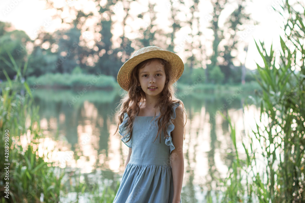 Cute girl in a blue dress and straw hat stands on the shore of the lake. Portrait of a child in the park with trees, green bulrush and a pond in summer. Young beautiful lady with curly hair on walking
