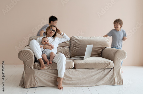 Young single mother working on laptop in loft sitting on couch while her sons running around her and shouting. High quality photo © natapetrovich