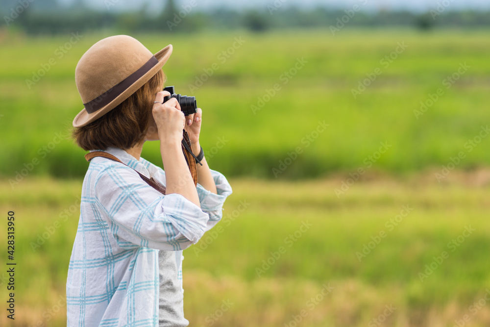 A tourist woman enjoy taking photo of landscape during her summer vacation holiday in the greenery field. A stylish traveler woman in hat taking photo of herself and landscape while relaxing A tourist woman enjoy taking photo of landscape during her summer vacation holiday in the greenery field. A stylish traveler woman in hat taking photo of herself and landscape while relaxing