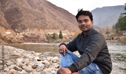 A happy man relaxing by mountain river enjoying natural landscape. Traveler backpacker sitting on rock on the Mangde chhu river bank in Bhutan kingdom. Summer trip photo