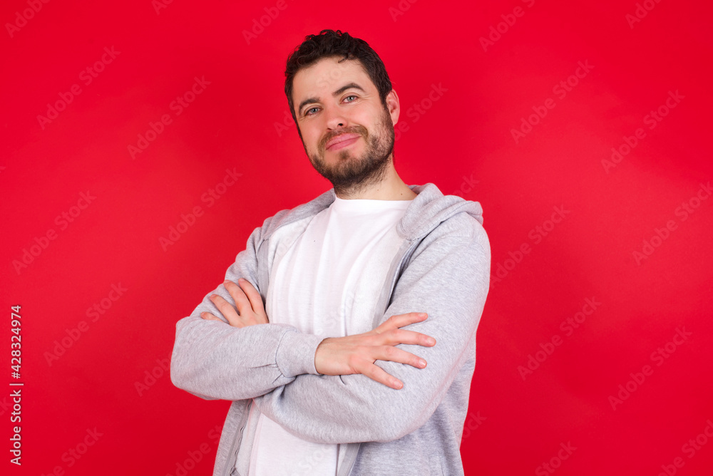 Waist up shot of self confident young handsome caucasian man in sports clothes against red wall has broad smile, crosses arms, happy to meet with colleagues. Waist up shot of self confident young handsome caucasian man in sports clothes against red wall has broad smile, crosses arms, happy to meet with colleagues.