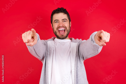 Close-up portrait of surprised young handsome caucasian man in sports clothes against red wall pointing with two fingers to the camera saying: I choose you!, looking up with open mouth. © Jihan