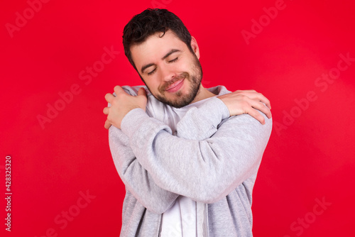 young handsome caucasian man in sports clothes against red wall. Hugging oneself happy and positive, smiling confident. Self love and self care.