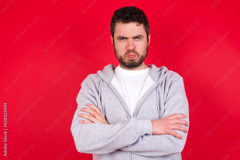 Gloomy dissatisfied young handsome caucasian man in sports clothes against red wall looks with miserable expression at camera from under forehead, makes unhappy grimace Gloomy dissatisfied young handsome caucasian man in sports clothes against red wall looks with miserable expression at camera from under forehead, makes unhappy grimace