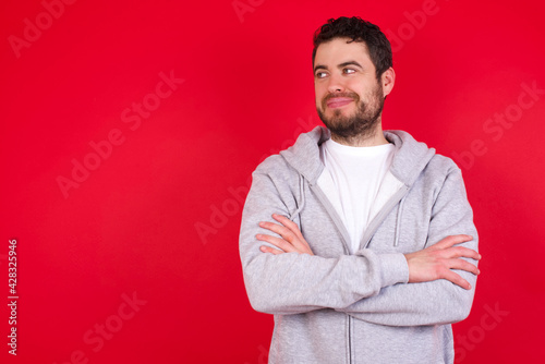 Pleased young handsome caucasian man in sports clothes against red wall keeps hands crossed over chest looks happily aside © Jihan